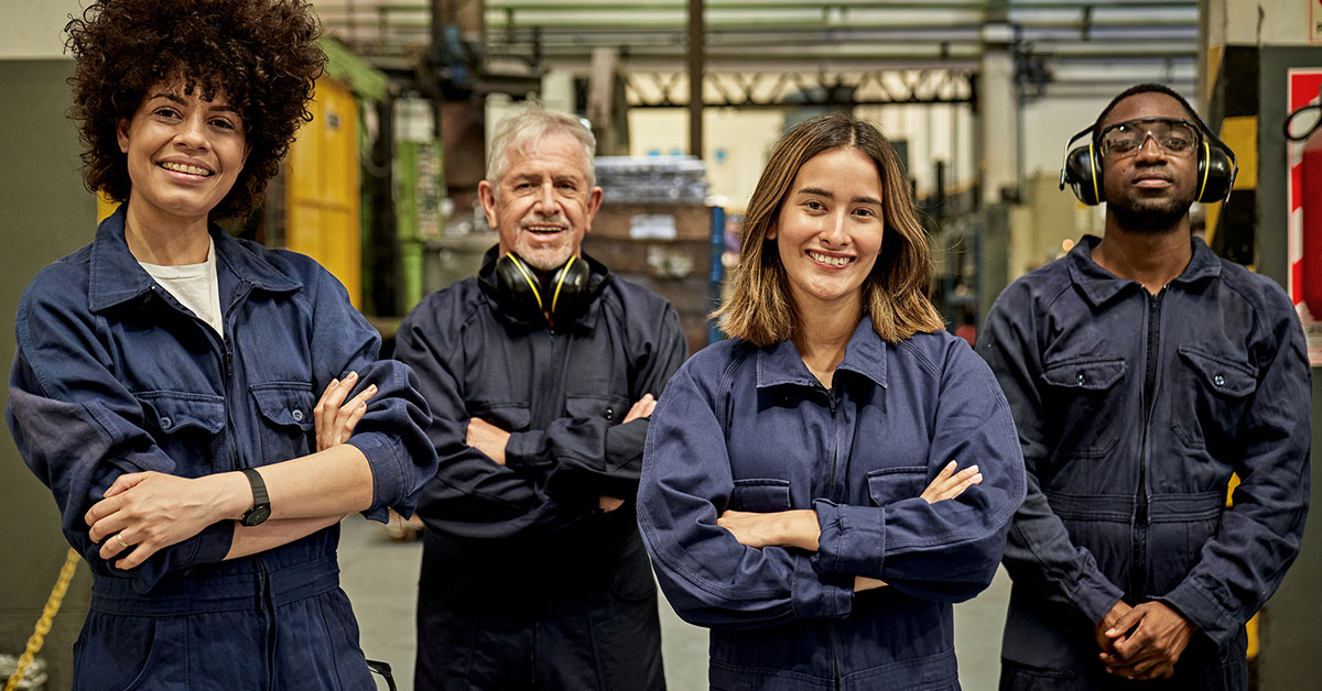Four male and female workers in coveralls, protective eyewear, and ear protectors standing with arms crossed and smiling at camera